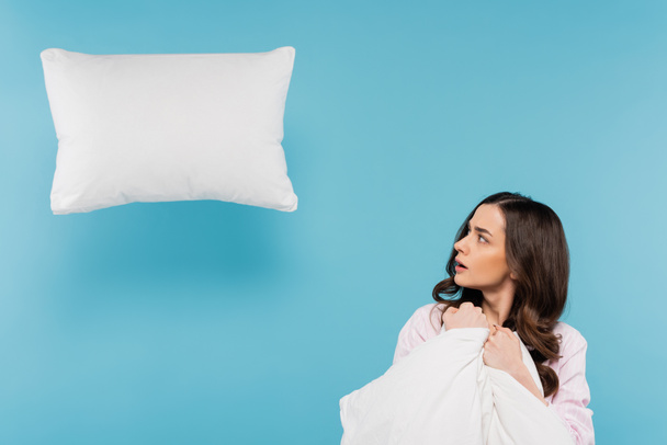 shocked woman in pajamas holding warm duvet looking at levitating pillow on blue  - Foto, Imagen