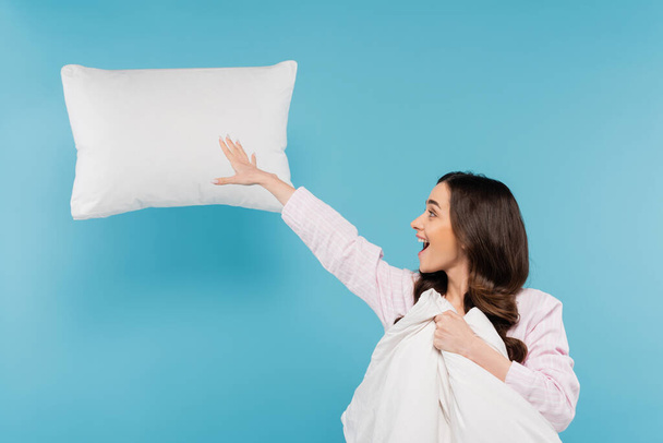 excited woman in pajamas holding warm duvet near flying pillow on blue  - Foto, Imagen