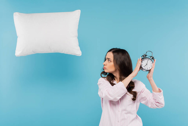 mujer joven molesta en pijama con reloj despertador vintage cerca de almohada voladora en azul  - Foto, imagen
