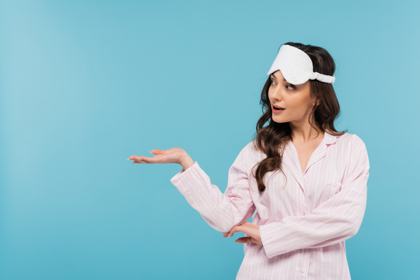 brunette young woman in night mask and pajamas looking away and pointing with hand isolated on blue  - Photo, Image
