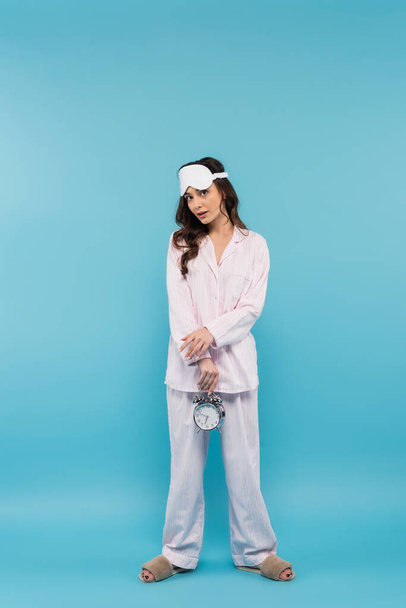 full length of young woman in night mask and sleepwear holding alarm clock on blue  - Foto, Imagen