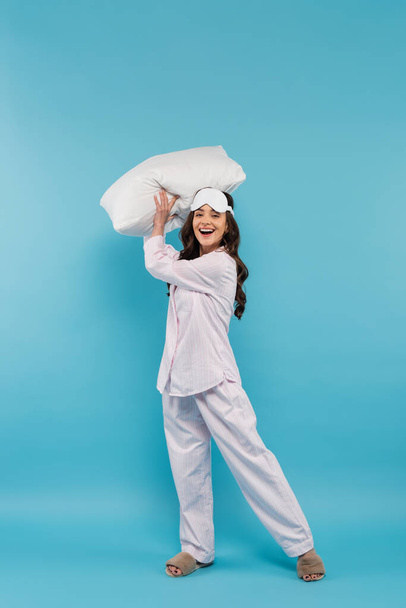 longitud completa de la mujer joven feliz en la máscara de noche y ropa de dormir con almohada en azul  - Foto, imagen