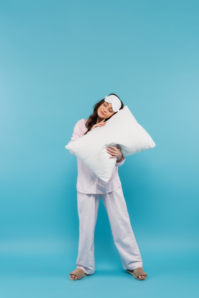 full length of sleepy young woman in night mask and pajamas holding pillow on blue  - Foto, imagen