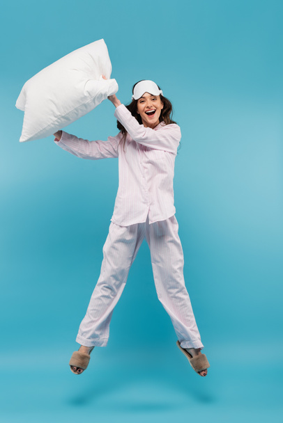 longitud completa de mujer joven sorprendida en máscara de noche y ropa de dormir con almohada y saltar sobre azul  - Foto, imagen