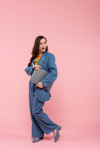 full length of amazed freelancer in blue pantsuit holding laptop on pink  - 写真・画像