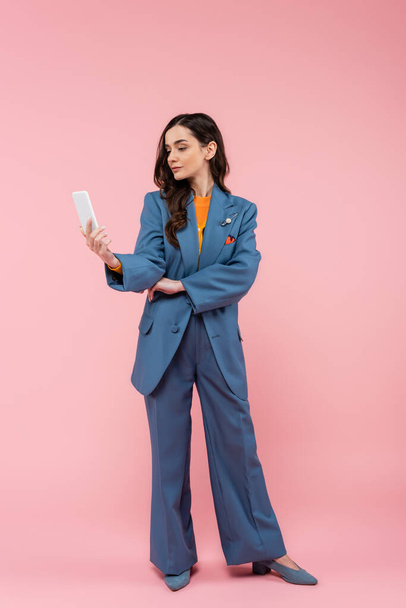 full length of young brunette woman in blue pantsuit using smartphone on pink  - Foto, Imagen