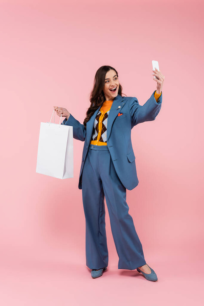 plná délka vzrušené ženy v modrých kalhotách drží nákupní tašku a přičemž selfie na růžové  - Fotografie, Obrázek