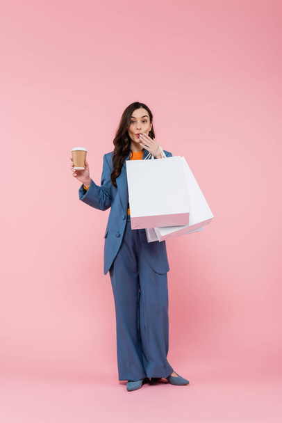 Полная длина эмоциональной женщины в синем брючном костюме держа сумки для покупок и кофе, чтобы пойти, прикрывая рот на розовый  - Фото, изображение