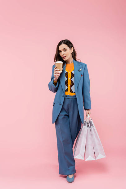 full length of brunette young woman in blue pantsuit holding shopping bags and coffee to go on pink  - Foto, afbeelding
