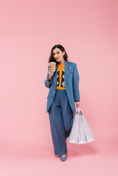 full length of brunette woman in blue suit holding shopping bags and coffee to go on pink  - Fotografie, Obrázek
