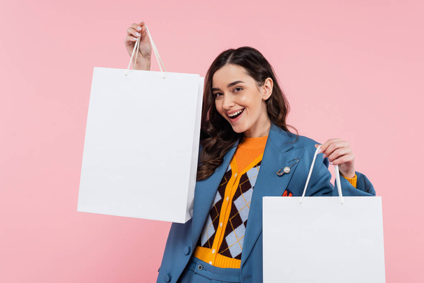 pleased young woman in blue blazer holding shopping bags isolated on pink  - Foto, Imagen