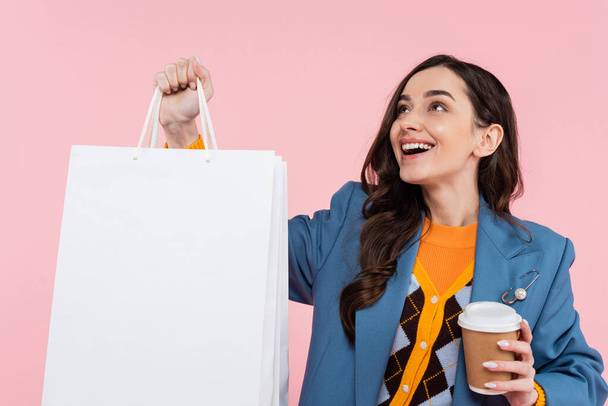 alegre joven en blazer azul sosteniendo bolsas de compras y café para ir aislado en rosa  - Foto, imagen