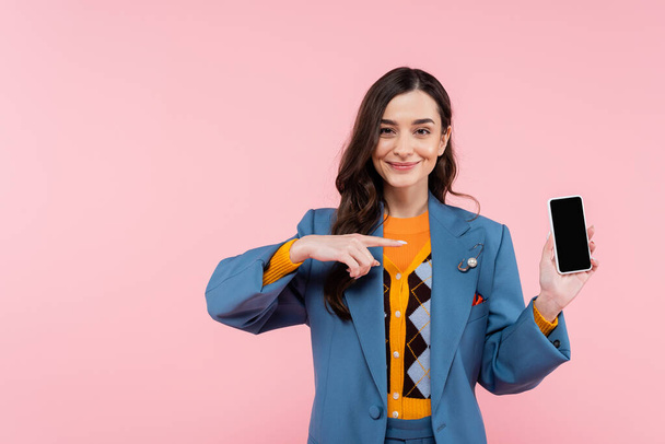 Mavi ceketli neşeli genç bir kadın pembe ekranda boş ekranlı akıllı telefonu işaret ediyor.  - Fotoğraf, Görsel