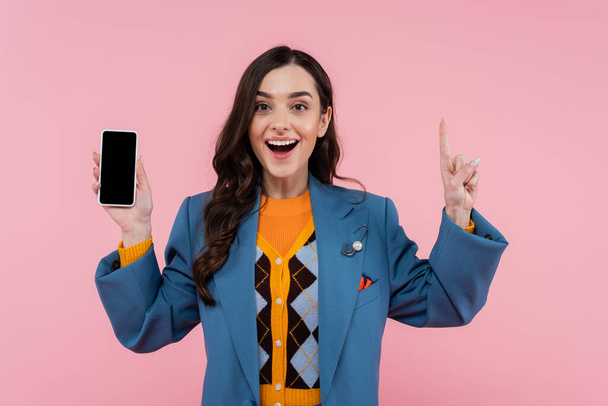 excited young woman in blue blazer holding smartphone with blank screen and pointing up isolated on pink  - Foto, Imagem
