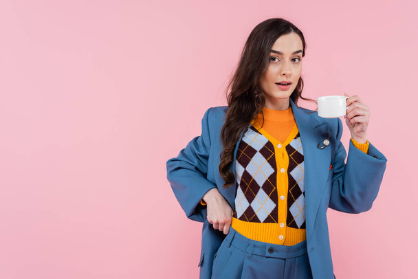 brunette woman in blue blazer holding cup of coffee isolated on pink  - Fotó, kép