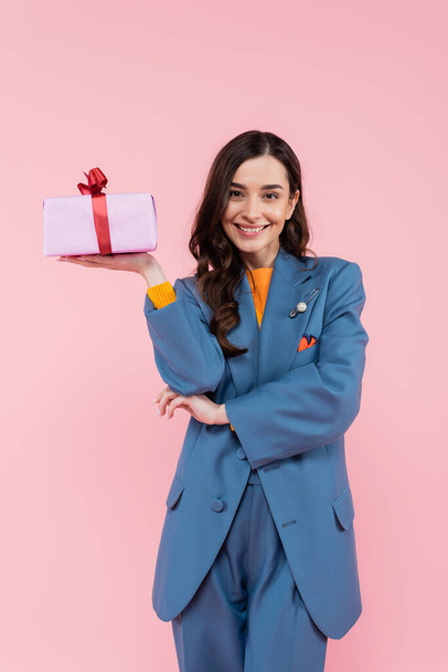 joyful young woman in blue blazer holding wrapped present isolated on pink  - Foto, Bild