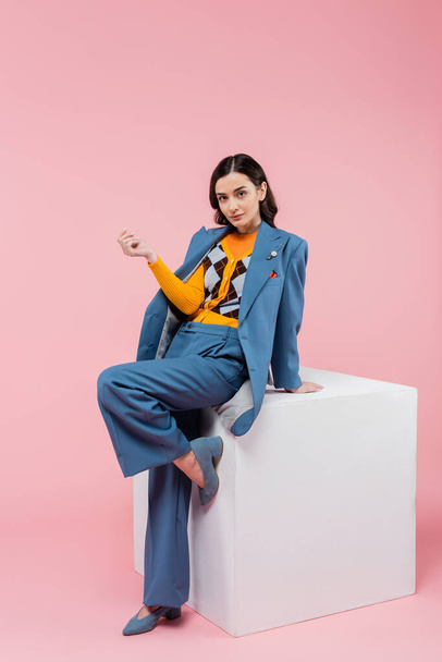 full length of trendy brunette woman in blue suit posing near white cube and looking at camera on pink background - Photo, Image