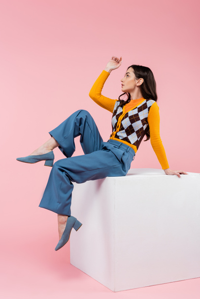 side view of young woman in stylish cardigan and blue pants looking away while sitting on white cube on pink background - Photo, image