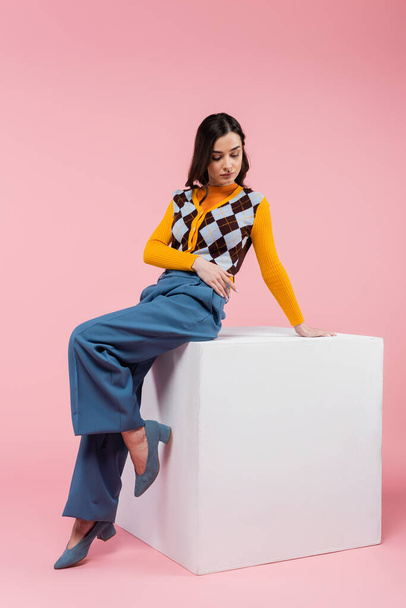 full length of brunette woman in blue pants and trendy cardigan sitting on white cube on pink background - Фото, изображение