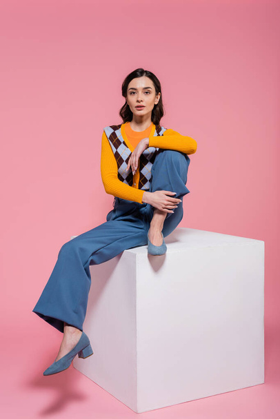 full length of woman in blue pants and trendy cardigan sitting on white cube and looking at camera on pink background - Photo, Image