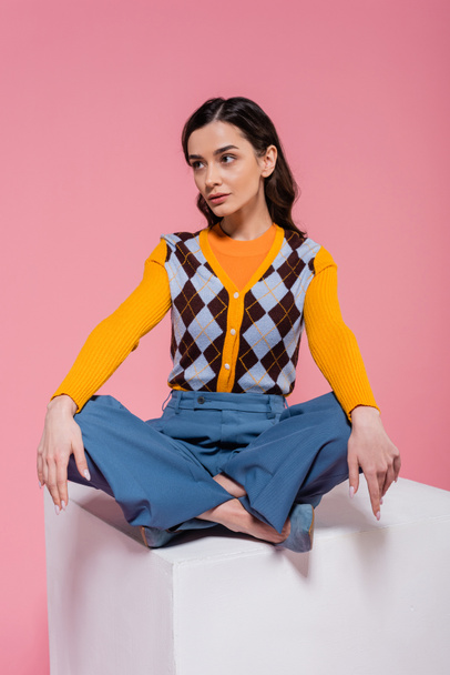 full length of brunette model in blue pants sitting with crossed legs on white cube on pink background - Zdjęcie, obraz