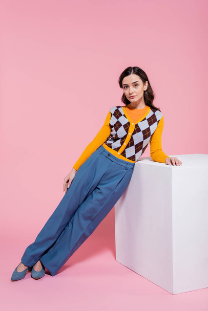 full length of trendy woman in blue trousers and orange cardigan with argyle pattern leaning on white cube on pink background - Fotoğraf, Görsel