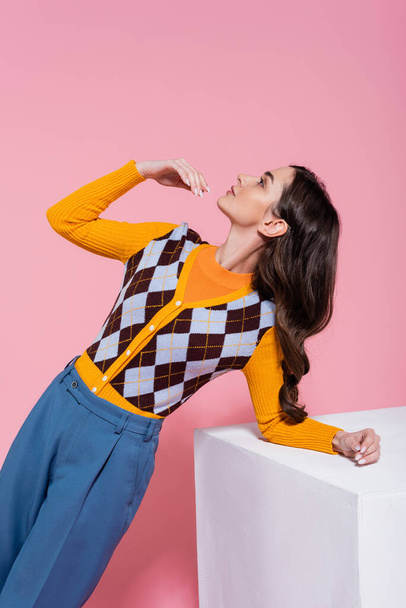 fashionable woman in bright cardigan and blue pants looking up while posing near white cube on pink background - Foto, afbeelding