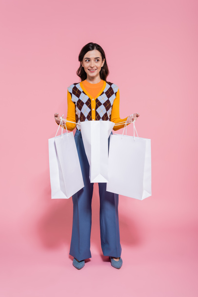 longitud completa de la mujer feliz en traje de moda sosteniendo bolsas de compras mientras mira hacia otro lado en el fondo rosa - Foto, Imagen