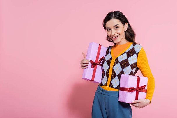 молодая женщина в ярком кардигане держит подарочные коробки и улыбается в камеру на розовом фоне - Фото, изображение