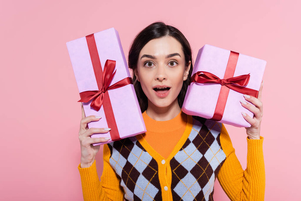 impressed woman in orange cardigan with argyle pattern showing gift boxes while looking at camera isolated on pink - 写真・画像