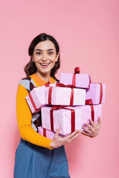 joyful brunette woman smiling at camera while holding pile of presents isolated on pink - Foto, Imagem