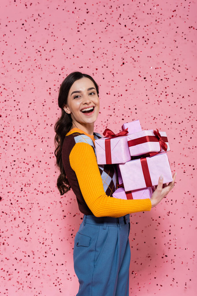 cheerful woman in stylish clothes holding gift boxes while looking at camera on pink background - Foto, Bild