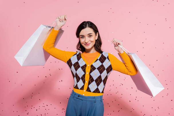 joyful woman in orange cardigan with argyle pattern posing with white shopping bags on pink background - Foto, afbeelding