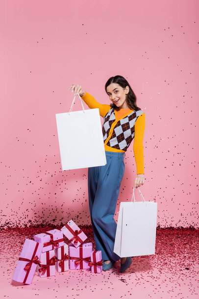 full length of fashionable woman posing with white shopping bags near gift boxes and festive confetti on pink background - Фото, изображение