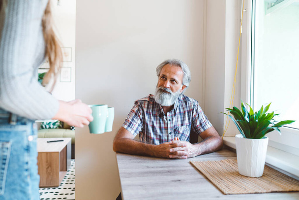 Young caucasian woman taking care of her grandfather, senior man with beard and grey hair. Granddaughter visiting her grandfather, spending quality time together. - Photo, Image