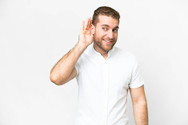 Young handsome blonde man over isolated white background listening to something by putting hand on the ear - Photo, Image