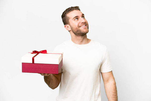 Joven guapo hombre rubio sosteniendo un regalo sobre fondo blanco aislado mirando hacia arriba mientras sonríe - Foto, Imagen