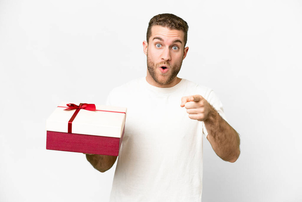 Young handsome blonde man holding a gift over isolated white background surprised and pointing front - Fotoğraf, Görsel