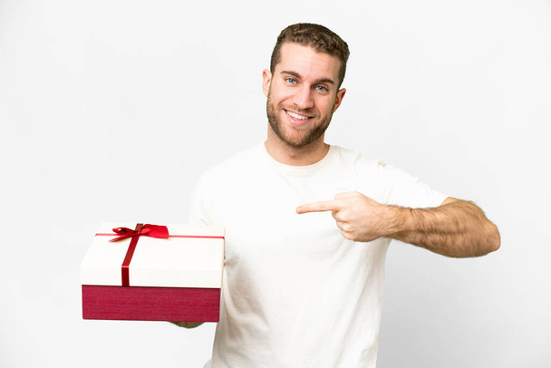 Fiatal jóképű szőke férfi kezében egy ajándék felett elszigetelt fehér háttér és rámutatva, hogy - Fotó, kép