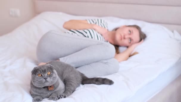 Depressed young woman lying in bed with a beautiful cat sitting next to her, unhappy woman suffering from insomnia or depression, psychological problem, blurred background - Felvétel, videó
