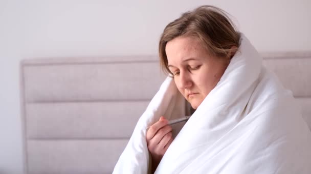 Portrait of sick depressed woman suffering from cold, measuring temperature with thermometer, sitting in bed covered with blanket at home. - Metraje, vídeo