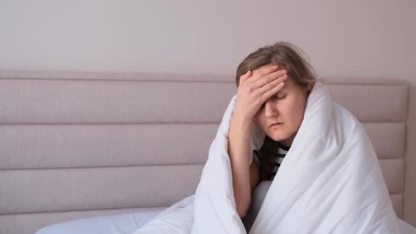 Άρρωστη νεαρή γυναίκα που κάθεται στο κρεβάτι και υποφέρει από πονοκέφαλο, κουρασμένη εξαντλημένη γυναίκα που καλύπτει το πρόσωπο με το χέρι τυλιγμένη σε ζεστή κουβέρτα. Βίντεο 4k - Πλάνα, βίντεο