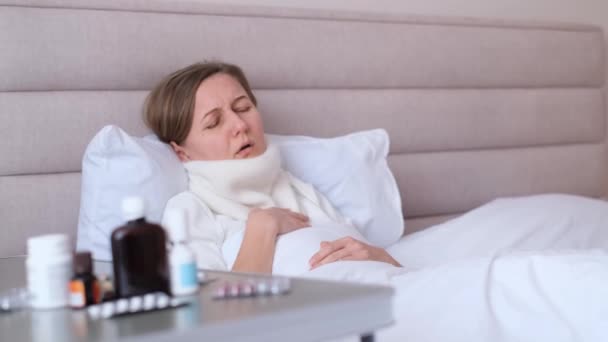 Tired sick girl with scarf around neck, covered with blanket, lying in bed and coughing, suffering from flu or cold and sore throat. Treatment of flu at home. - Metraje, vídeo