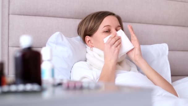 Een jonge vrouw niest in een tissue op een wit bed in de slaapkamer, griep en allergie concept ziek meisje met griepsymptomen hoesten thuis. - Video
