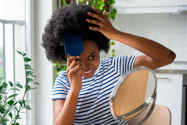 Cool African woman with comb sticking out of curly hair smiling looks in mirror and touches wig with hands. Positive black teen girl with wavy hairstyle grooms herself sits at table in home - Photo, Image