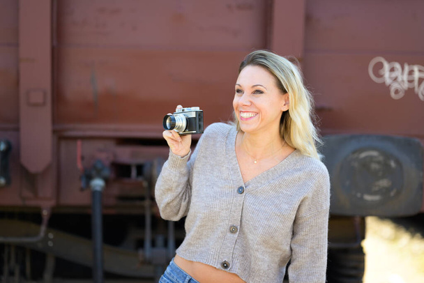 Zijaanzicht van een enthousiaste glimlachende vrouw van middelbare leeftijd met een oude camera in een bruine trui voor een oude treinwagon - Foto, afbeelding