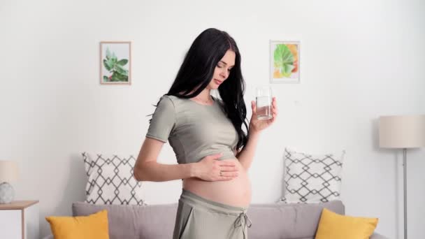 молодая беременная женщина нежно касается живота и пьет здоровую чистую воду из стекла для поддержания водного баланса в организме, смотрит в камеру - Кадры, видео