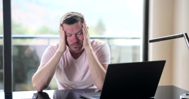 Un homme fatigué à la maison devant un ordinateur portable se masse la tête, au ralenti. Travail à distance, situation stressante - Séquence, vidéo