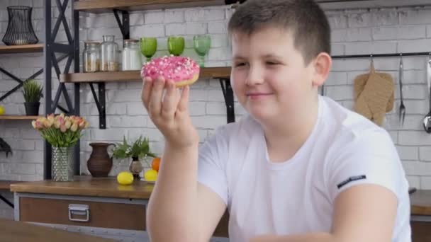 Teenager eats junk food while working at home. Attractive plump handsome young guy feels happy and enjoys eating a donut in the kitchen at home. 4k - Séquence, vidéo