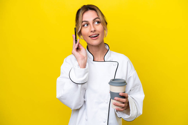 Уругвайская шеф-повар женщина изолированы на желтом фоне держа кофе, чтобы забрать и мобильный - Фото, изображение